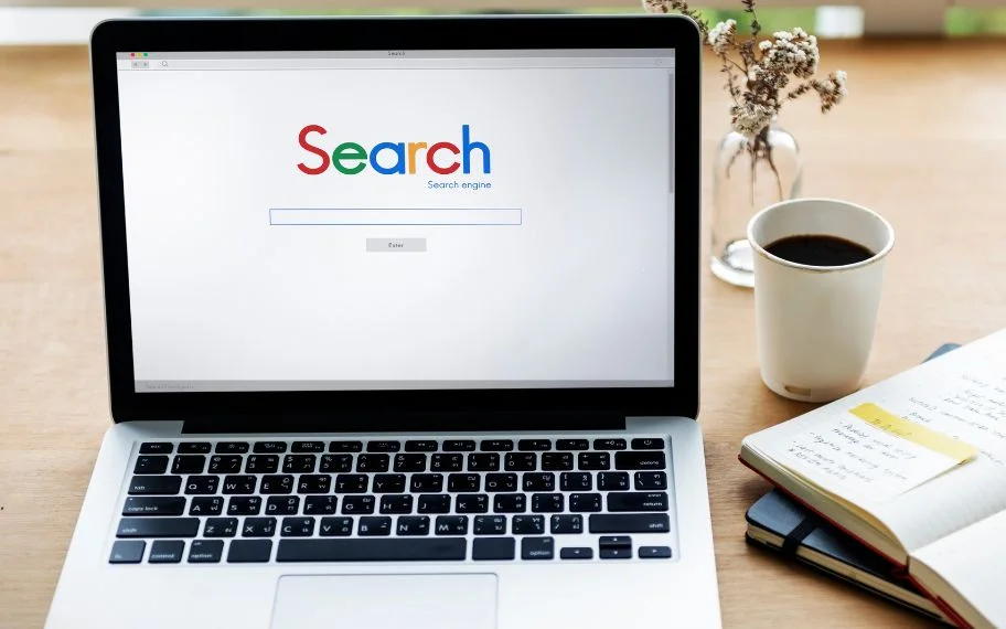 Laptop on a desk showing a search engine homepage, next to a cup of coffee and an open notebook.