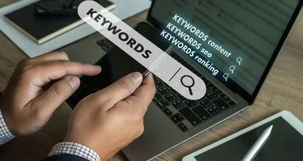Hands holding a keywords search bar in front of a laptop listing keyword content, SEO, and ranking searches.